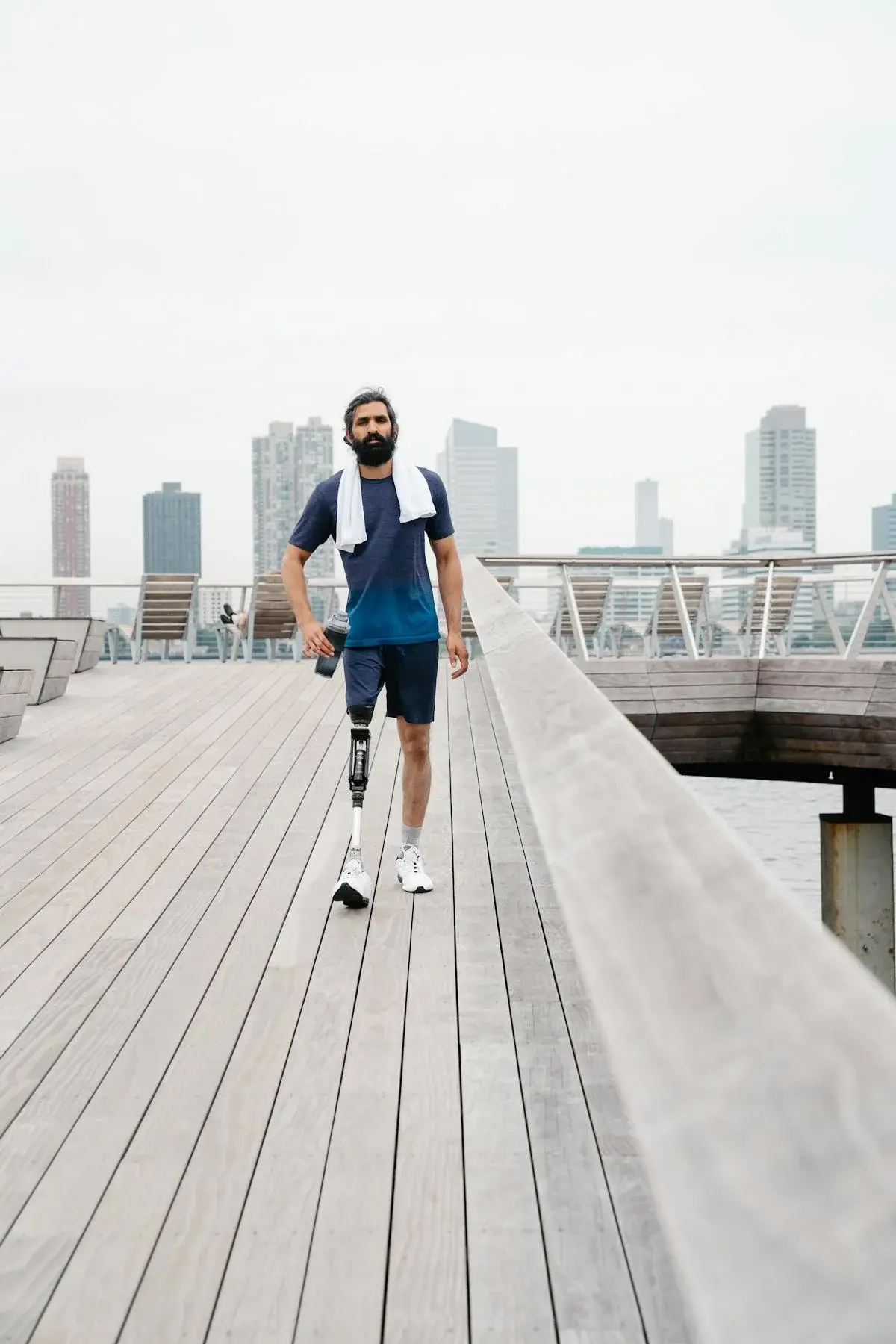A man with a below-knee prosthetic leg walks confidently along a waterfront boardwalk, water bottle in hand